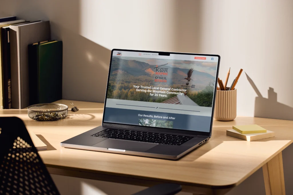 Laptop on a modern wooden desk displaying a custom website for Kor Roofing & O’Neil Restoration, showcasing their contractor services with a scenic mountain background.